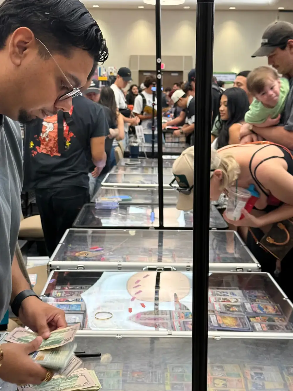 Cash deals at The Cactus TCG Show vendor hall Vendor counting cash across glass cases of graded Pokémon cards while families browse The Cactus TCG Show in Mesa, Arizona
